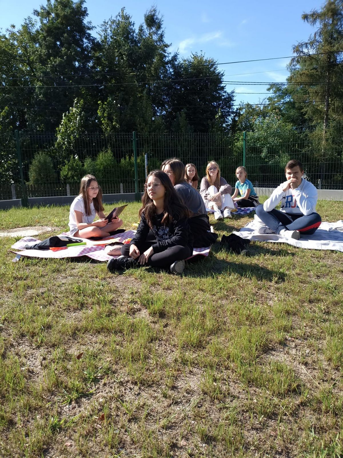 Uczniowie siedzący przed szkołą na kocach 